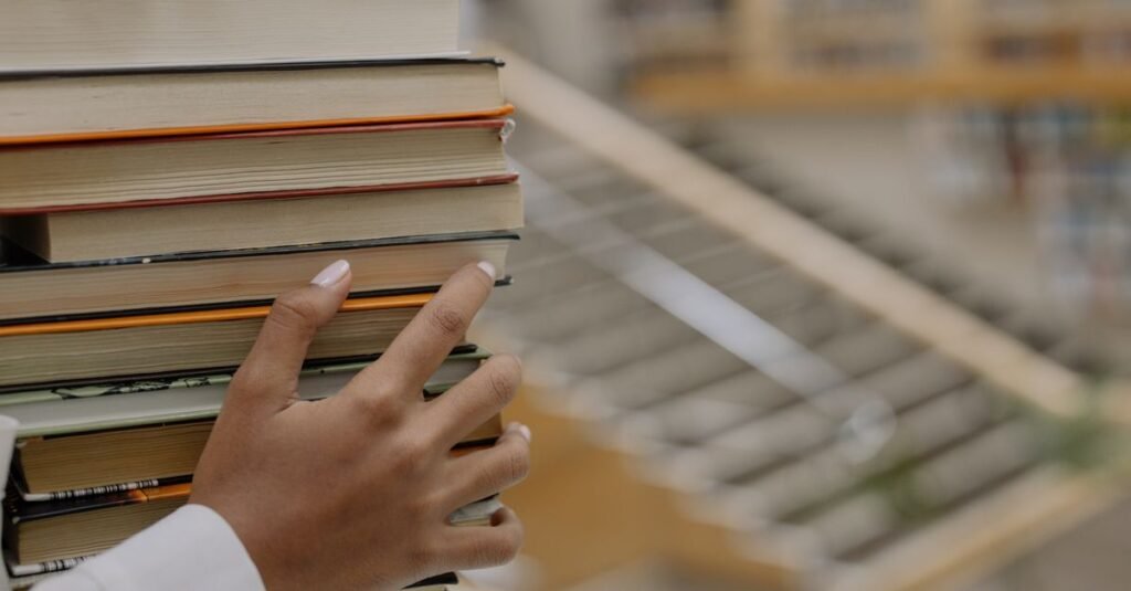 Hand holding a stack of books with library shelves blurred behind